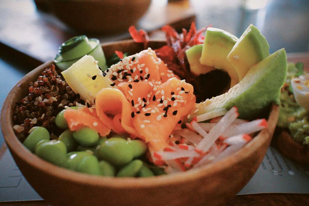 Nahaufnahme einer bunten Poké-Bowl mit Lachs, Avocado, Edamame, Quinoa und Sesam in einer Holzschale.