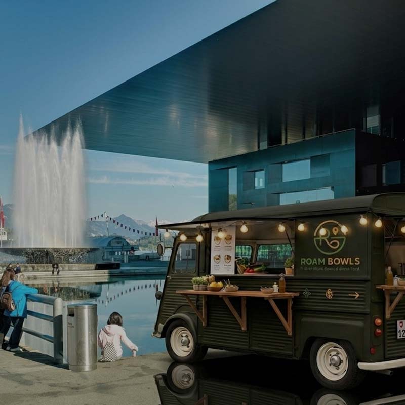 Der dunkelgrüne Roam Bowls Foodtruck steht vor dem modernen KKL Luzern direkt am Seeufer, im Hintergrund ist eine grosse Wasserfontäne zu sehen.