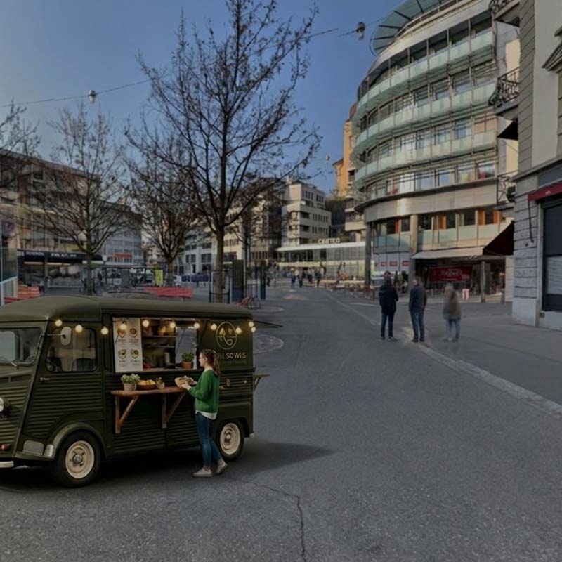 Ein dunkelgrüner Foodtruck steht beim Löwenplatz vor modernen Gebäuden, während eine Kundin am Tresen bedient wird.