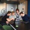 Vier gut gelaunte Teammitglieder sitzen in einem modernen Büro um einen Tisch und arbeiten gemeinsam an Laptops.